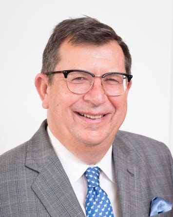 The Hon A S Bell smiling. He wears glasses, a grey suit, blue tie and white shirt.