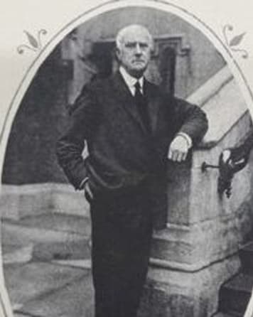 Black and white photograph of a man in a suit with tie. He is leaning on a stone staircase banister.