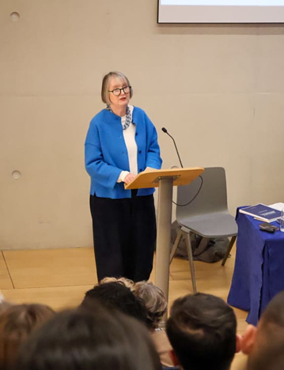 A woman with glasses and a blue jacket speaks at a podium in a lecture room. The audience listens intently, creating an atmosphere of intellectual engagement.