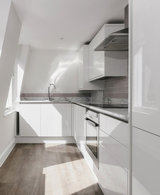 Modern kitchen with glossy white cabinets, grey subway tile backsplash, and wood flooring. Stainless steel appliances, including an oven and sleek sink, are integrated. Sunlight streams through a window, enhancing the clean, minimalist aesthetic.