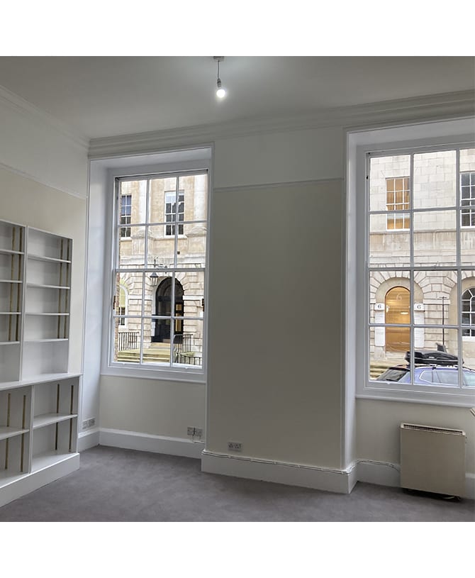 A bright, empty room features two large windows with white frames, overlooking a historic stone building. There are white shelves on one wall.