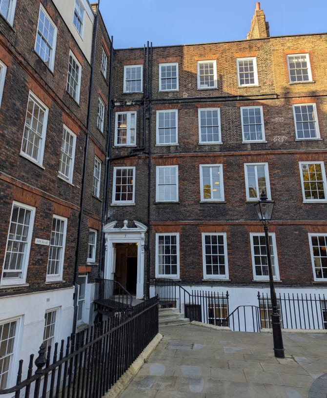 Historic brick building with large white-framed windows under a clear blue sky. Wrought iron railings and a lamppost line the stone pathway.