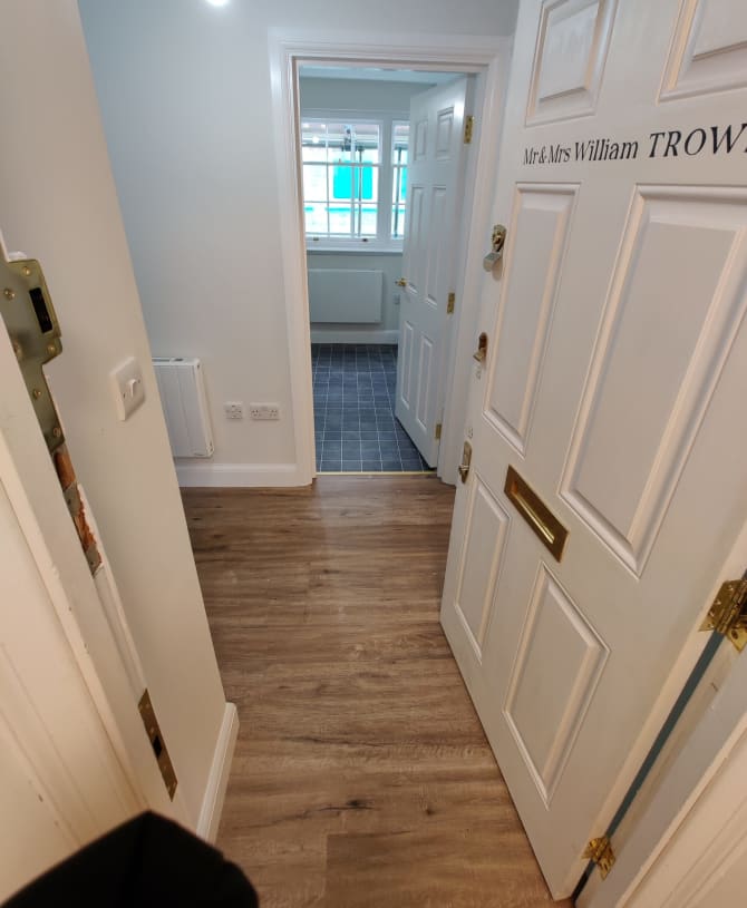 A bright hallway with wooden flooring leads to an interior room with dark tile flooring. An open door displays a nameplate. Light from a bulb illuminates the space.