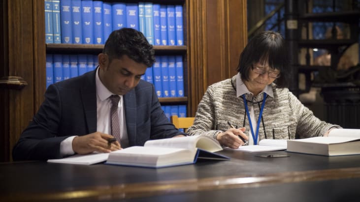 Two visitors sit at a desk at Lincoln's Inn Library, making notes in their exercise books. Books from the Library lie open on the desk in front of them. The visitor on the left (facing the camera) wears a navy, chequered suit, a white shirt and a tie with a black and red pattern. The visitor on the right wears a cardigan over a white blouse.