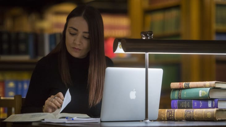 A women studying with books and a laptop in the Inn Library, a desk lamp in front of her