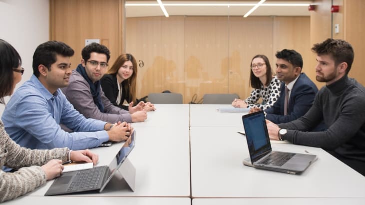 A meeting takes place in the Ashworth Centre. Attendees sit around rectangular tables pushed together in a boardroom format. A laptop and a tablet with a keyboard attached are open on the desks.