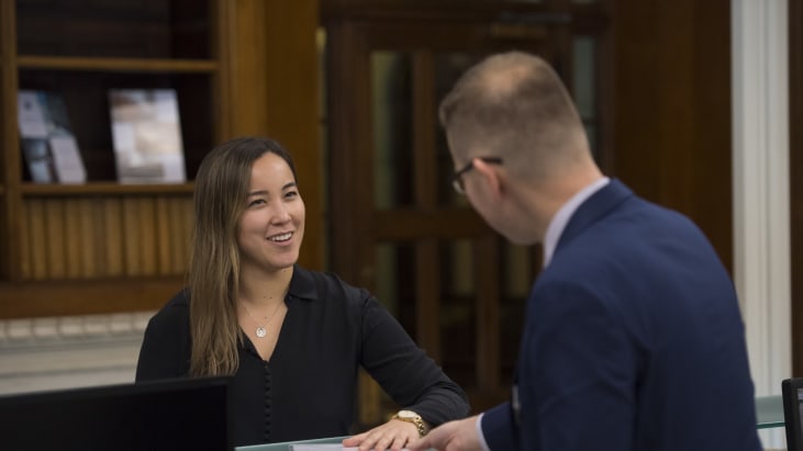 A smiling woman places her hand on a document at the reception desk of Lincoln's Inn. A member of staff, blurred in the foreground, reaches to pick it up.