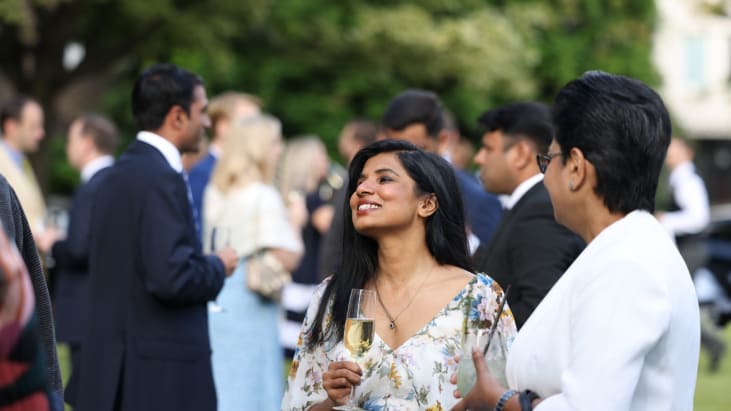 A guest smiles away from camera on the North Lawn of Lincoln's Inn, stood away from a large crowd, who are out of focus in the background. She wears a floral dress and holds a glass of champagne in her hand.