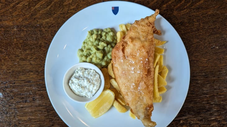 Fish and chips, laid on a plate served with mushy peas, a wedge of lemon and tartare sauce in a small container.