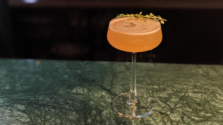 A peach coloured cocktail in a champagne coupe with a sprig of thyme for garish