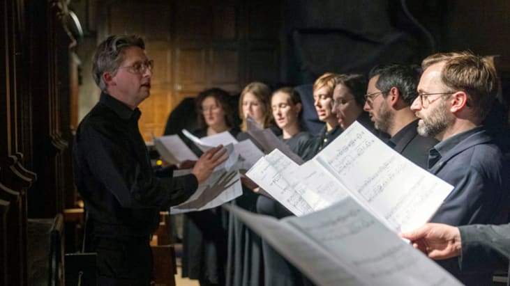 The choir performing in the Great Hall minstrel gallery. They hold music, are dressed in black and are being conducted by Nicholas Shaw