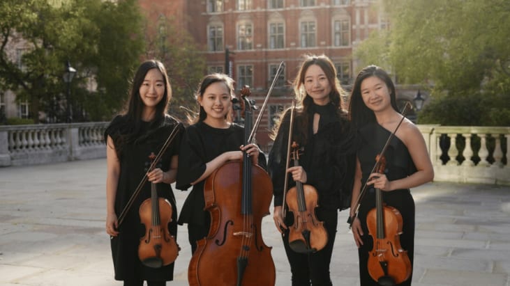 Four smiling women in black outfits stand together outdoors, each holding an upright cello against a city-building backdrop, conveying a warm, confident musical ensemble.