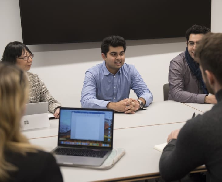 A group of people sit around a grey conference table. A Mac is open in the foreground.