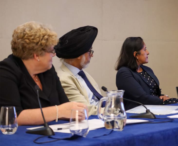 Chief Master Shuman, The Rt Hon. Lord Justice Singh and The Hon. Mrs Justice Cheema-Grubb, look at The Hon Mr Justice Choudhury as he answers a question. They are sat behind a table with water jug, papers and microphones on it, the table has a blue table cloth.