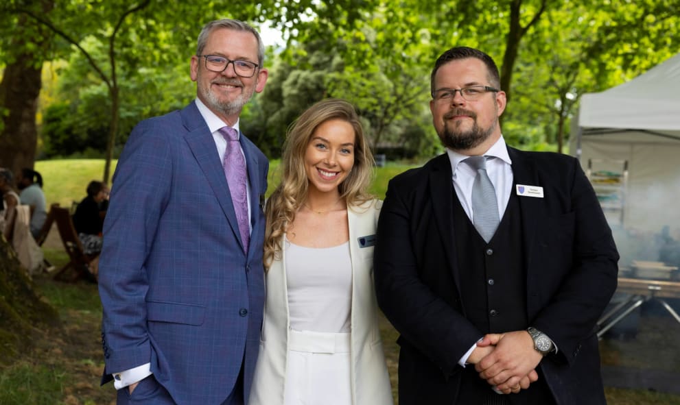 Three people stood in a garden. Two men in dark coloured suits and a woman stood in-between the men dressed in a white suit