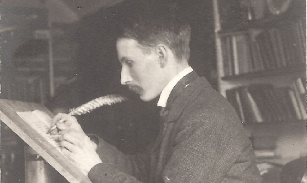 Black and white photograph of Edward Johnston writing with a feathered quill on a raised writing desk. He is a white man with a prominent moustache.