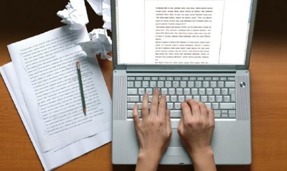 hands on laptop with papers at side - image representing essay writing