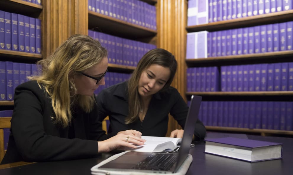 One woman helps another with her research in the Library of Lincoln's Inn, shelves of blue leather bound books behind them. They sit at a desk with a laptop in, but are looking through paper notes