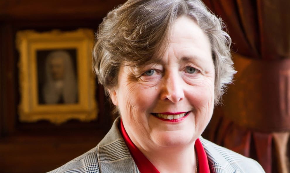 Susanna McGibbon KC (Hons) smiling. She wears a grey jacket, red blouse and red lipstick and has short gray hair. The wall behind her is dark wood panelling.