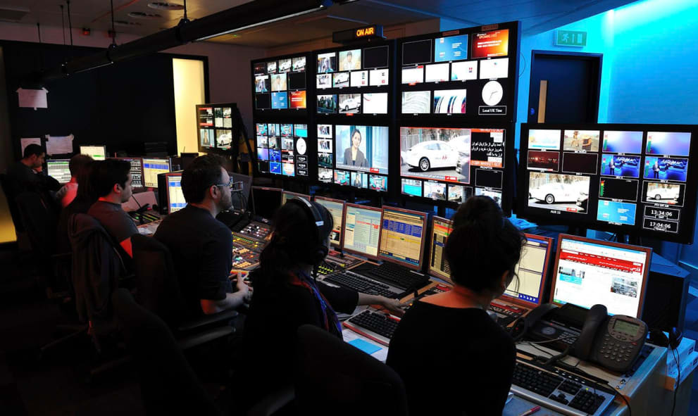Three people looking at a TV gallery in BBC News. There are dozens of TVs in a dimly lit room.