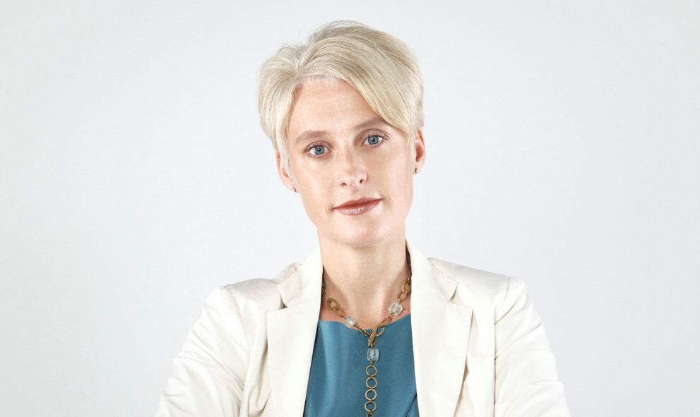 Headshot ofEmma Hopkins OBE. She wears a white suit jacket, teal top and gold jewelry and has short blonde hair.
