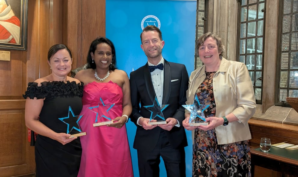 4 winners at the Employed Bar Awards hold their glass trophies and smile in front of a Bar Council branded banner