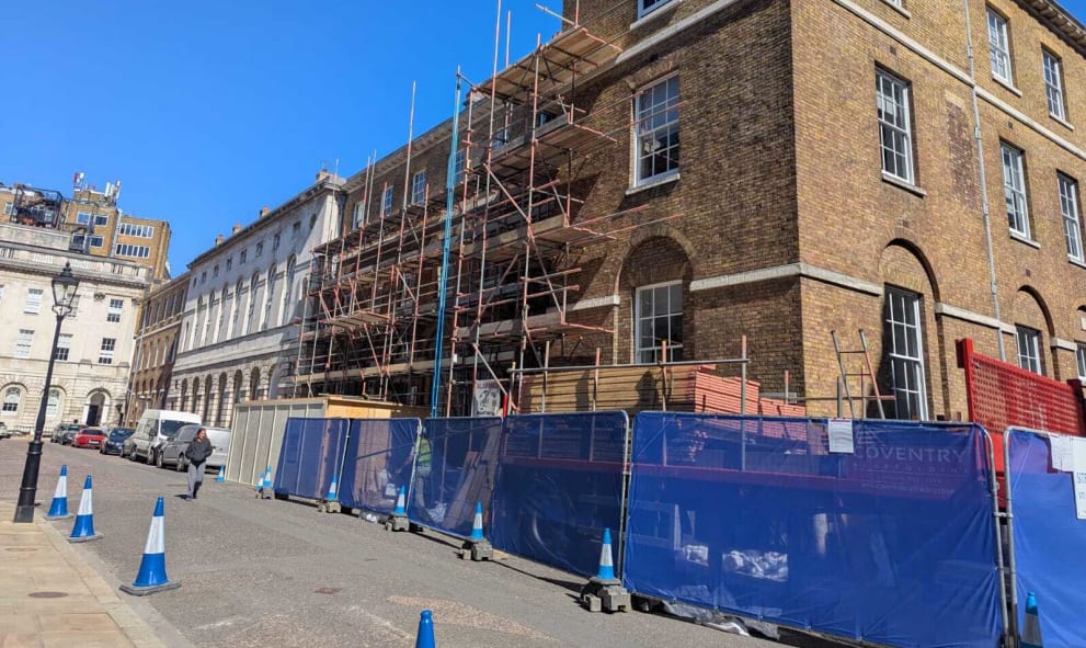 Historic brick building (8 & 9 Stone Buildings) under renovation on a sunny day. Scaffolding covers the facade. Blue barriers and cones line the street, ensuring safety.