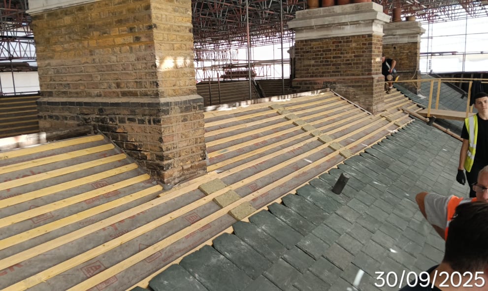Scaffolding surrounds a brick building under renovation. Workers in hi-vis vests are present on the unfinished roof. Date in corner: 30/09/2025.