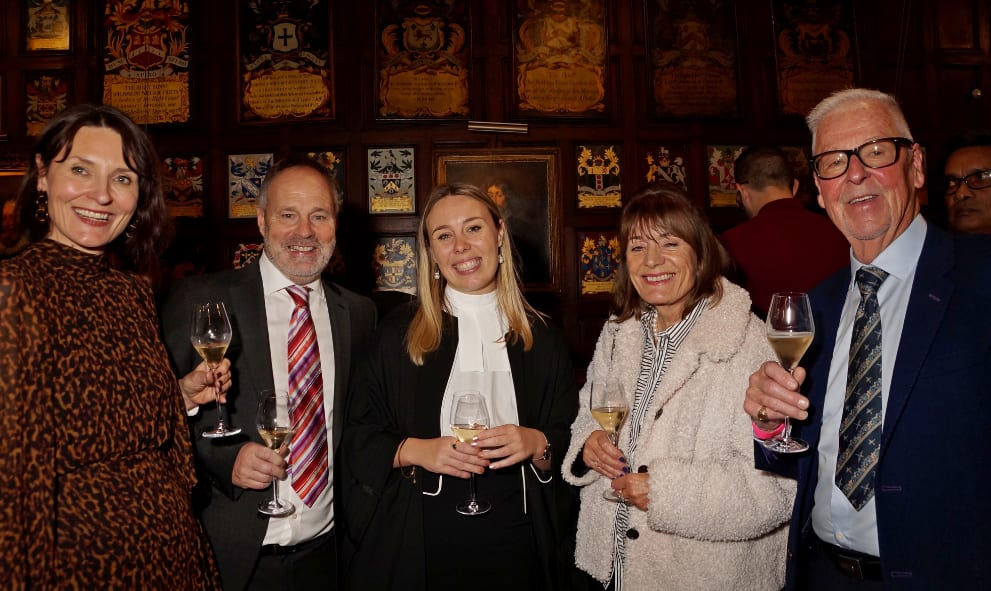 A group of smiling people holding champagne glasses, standing in a warmly lit room with ornate wooden panels and plaques on the walls, conveying a celebratory mood.