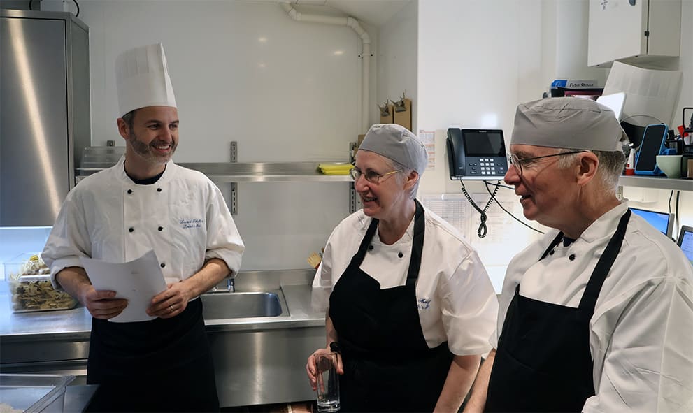 The Treasurer (The Hon Sir Paul Morgan) and his wife in the pastry room ofthe kitchen, in chefs whits, with pastry chef Laurent showing them around