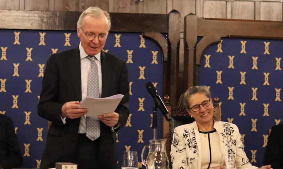 A man in a suit speaks at a podium, holding a paper. A woman in a patterned blazer sits smiling beside him. The setting is formal with a blue patterned backdrop.
