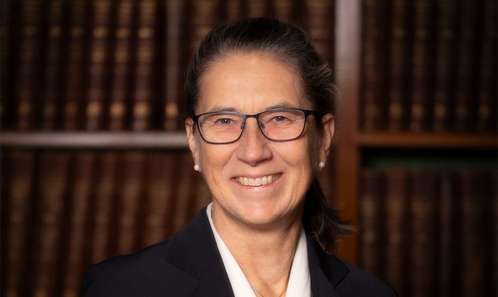 Elspeth Talbot Rice KC, smiling, in front of a dark wooden book shelf of old leather bound books. She wears a white blouse, black suit jacket, pearl earrings and glasses. He brown hair is tied back in a low ponytail.