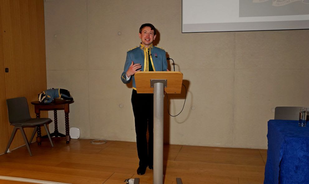 A person in a blue military-style uniform speaks at a podium in a modern conference room. They appear confident and engaging. To the left is a small table and chair, and to the right, a blue-covered table.