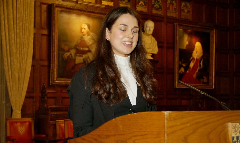 Woman with long hair speaking at a wooden podium in a room decorated with classical paintings and busts.