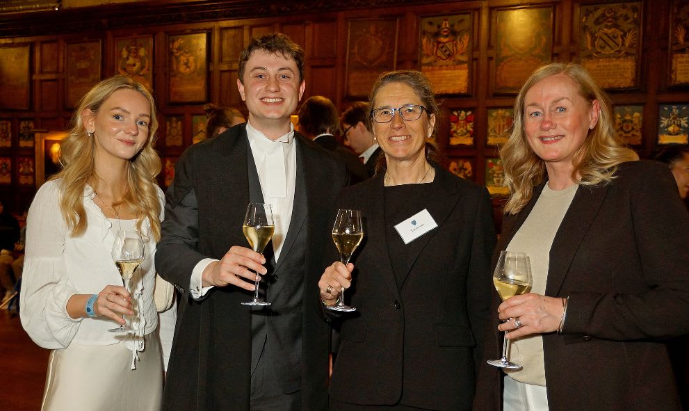 Four elegantly dressed people holding wine glasses at a formal indoor event with ornate wooden paneling in the background.