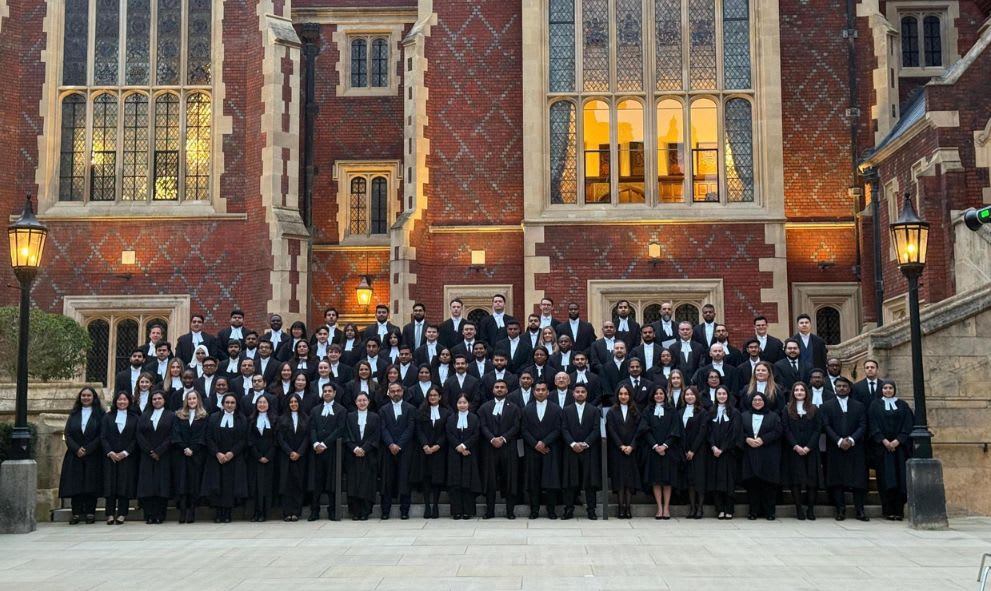 Large group of graduates in black gowns posing on steps of a historic brick building with lit windows at dusk.