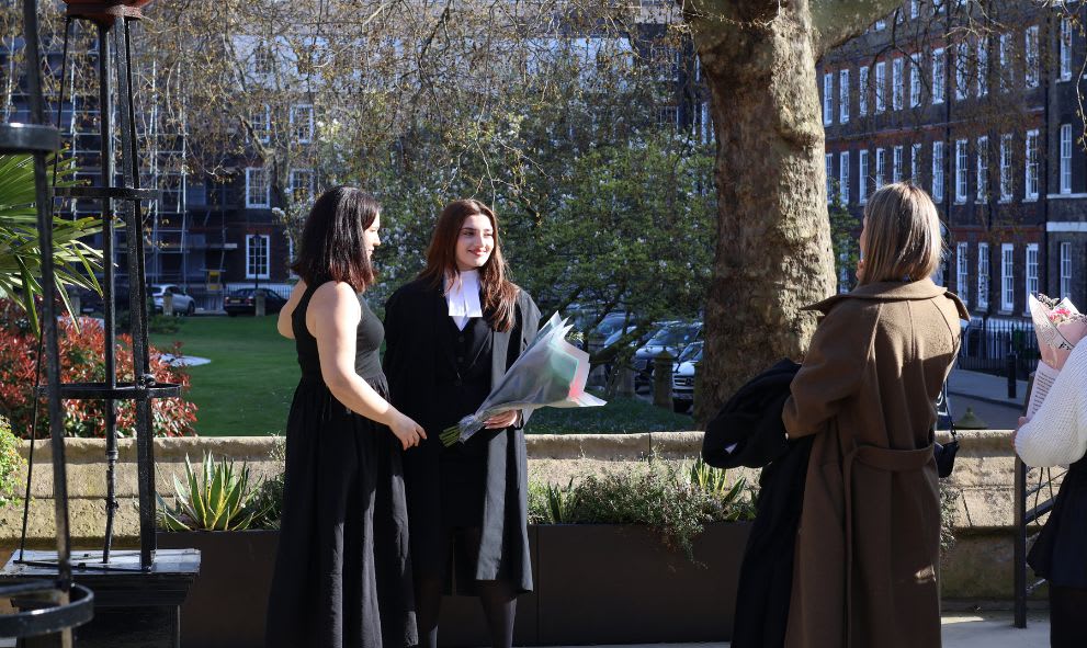 Two women in formal attire stand outdoors, one holding a bouquet, while two others observe nearby.