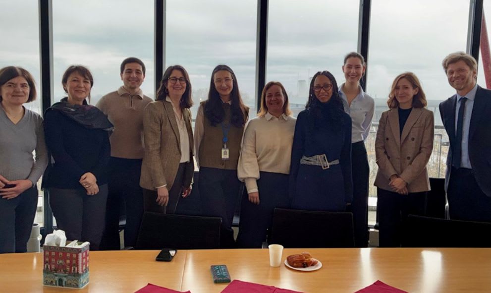 A group of ten people stands smiling in front of large windows. A table with tissues, a smartphone, cup, and pastries is in the foreground. Bright, casual, and friendly atmosphere.