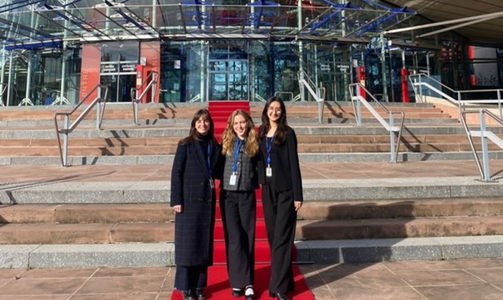 Three people stand on steps outside a modern glass building with a red carpet. They are smiling and wearing ID badges. The mood is cheerful.
