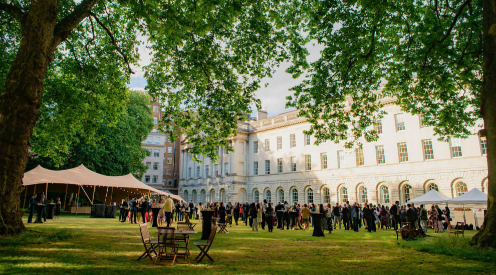 A photo of people at garden party in manicured lawns with a back drop of a white building and a stretch tent in the background. People are eating canapes and drinking bubbles.