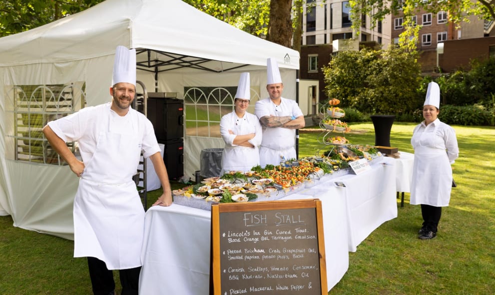Chefs stood next to a fish food stall, All chefs wearing white chef overalls with a tall chef hat