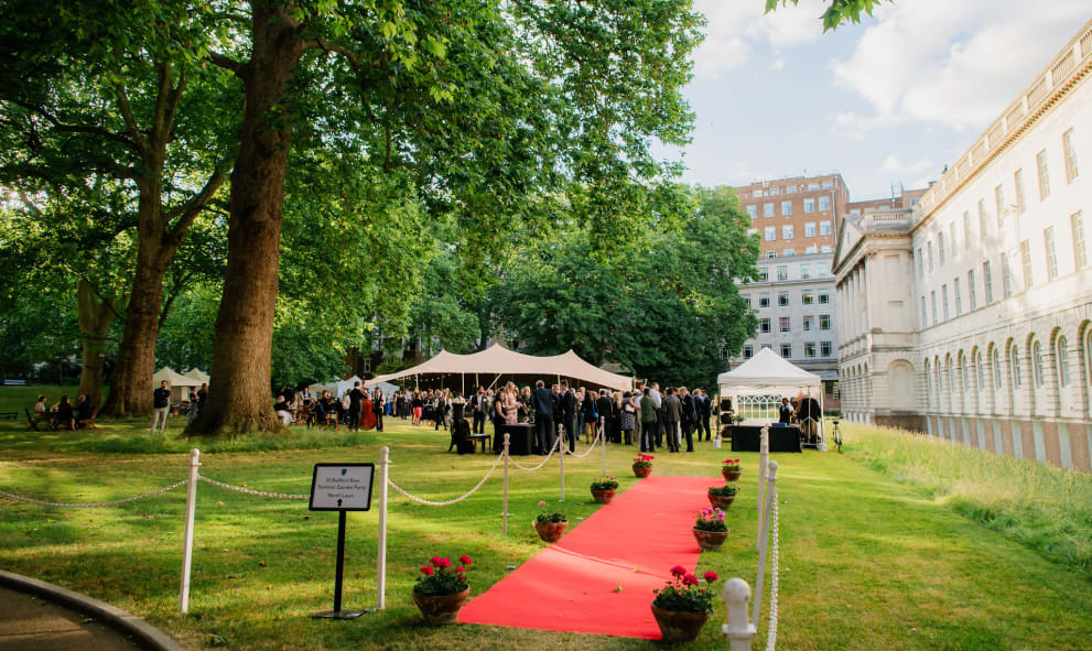 A photo of a red carpet on a lawn leading to people stood at a drinks reception in a garden on a sunny day.