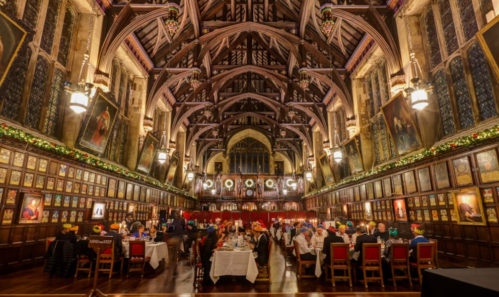 Wide shot of guests eating christmas dinner in the Great Hall, wearing crack crown. The Hall is decorated with garlands and wreaths lit with fairy lights