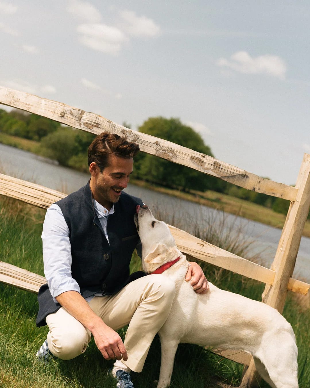 Man in a navy Darzi gilet crouching by a wooden fence with a yellow Labrador