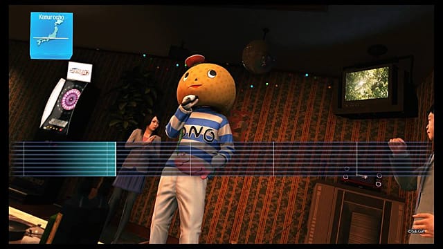 A person stands on a stage wearing an orange mascot head while singing karaoke