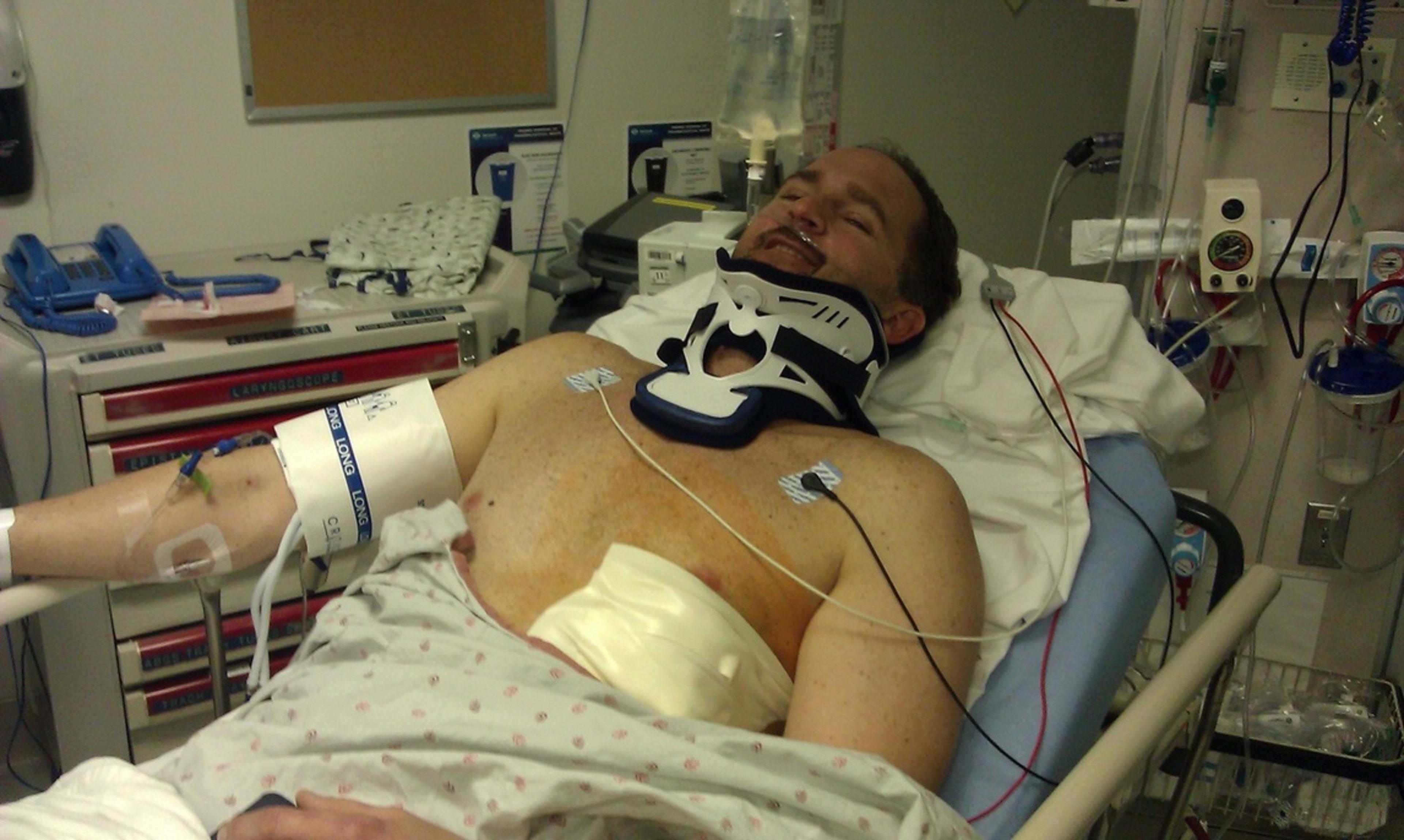 Bill in a hospital bed, neck brace and monitors, and still smiling.