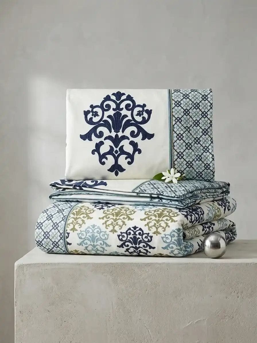 A folded bedding set with a white base and ornate navy blue damask-style patterns, stacked on a light stone pedestal against a neutral gray wall. A small white flower and a silver decorative sphere rest beside the stack.
