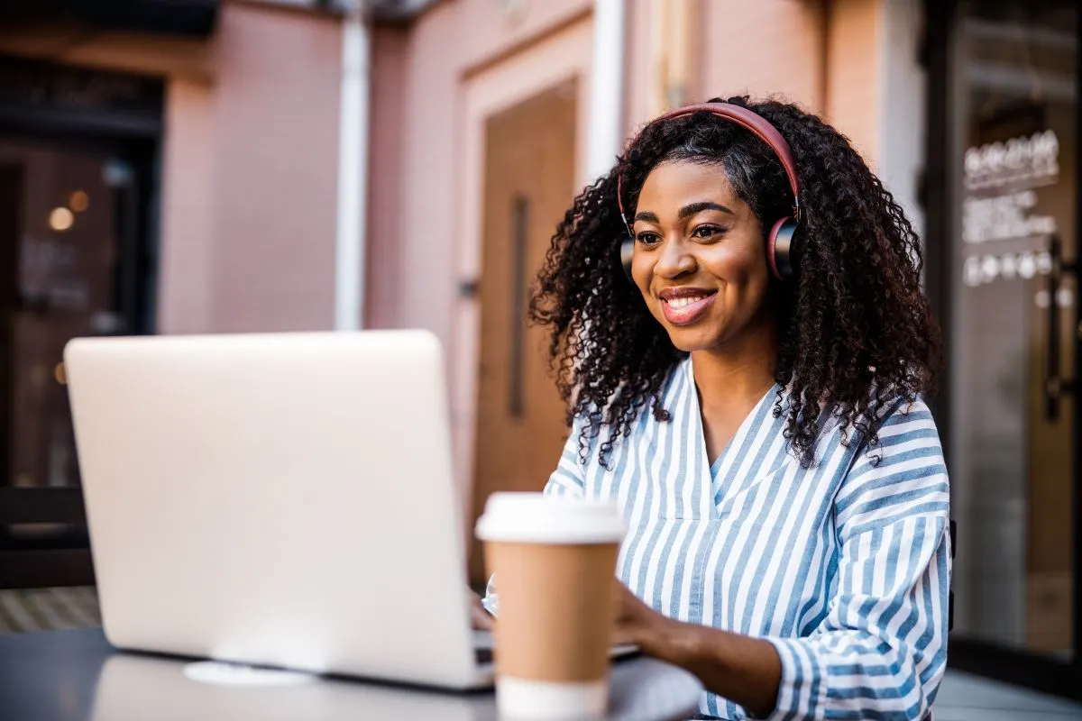 A woman wearing headphones and a striped shirt is smiling while using her laptop at an outdoor café table with a takeout coffee cup, possibly analyzing Google Ads KPI data.