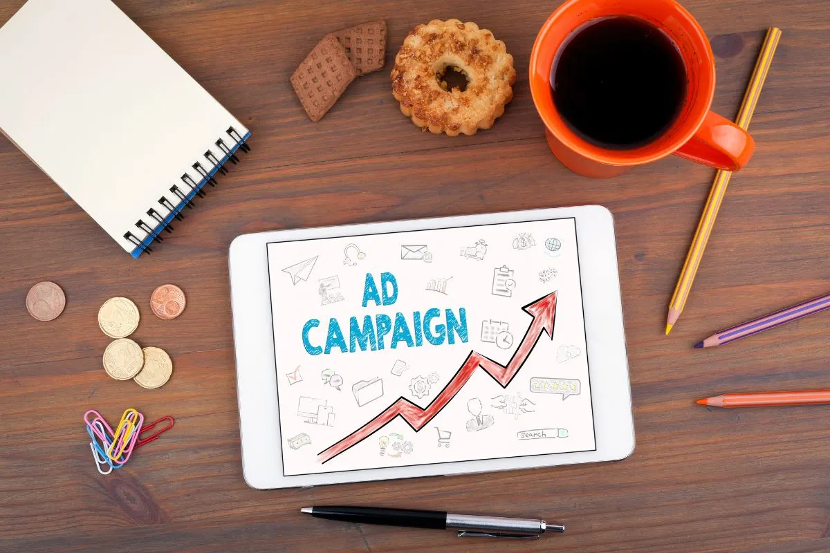 A Tablet On A Desk Displays A Graph Titled &Quot;Ad Campaign,&Quot; Comparing Google Ads Vs. Facebook Ads, Surrounded By A Notepad, Coins, Cookies, A Cup Of Coffee, And Pencils.
