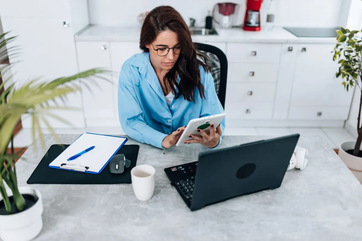A Woman In A Blue Shirt Sits At A Table With A Laptop, Clipboard, And Coffee Cup, Holding A Tablet In Her Bright Kitchen As She Researches How To Optimize Her Google Business Profile.